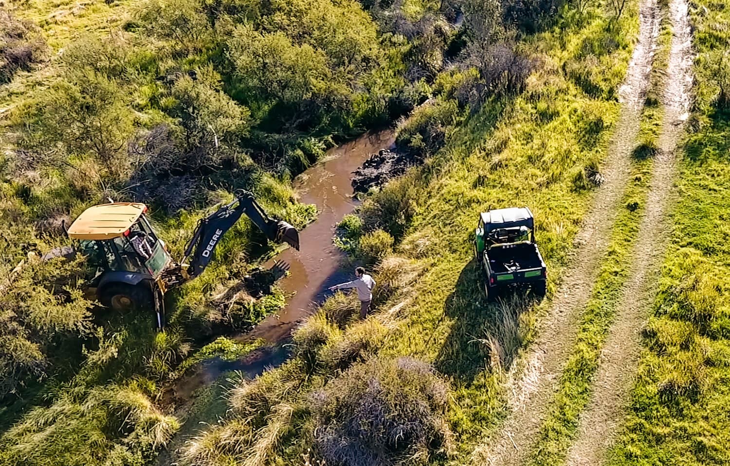 Vista aérea de una retroexcavadora y un vehículo de apoyo junto a un arroyo